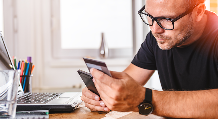 A man is holding a smartphone in one hand and a credit card in the other. He is typing his credit card number into his phone.